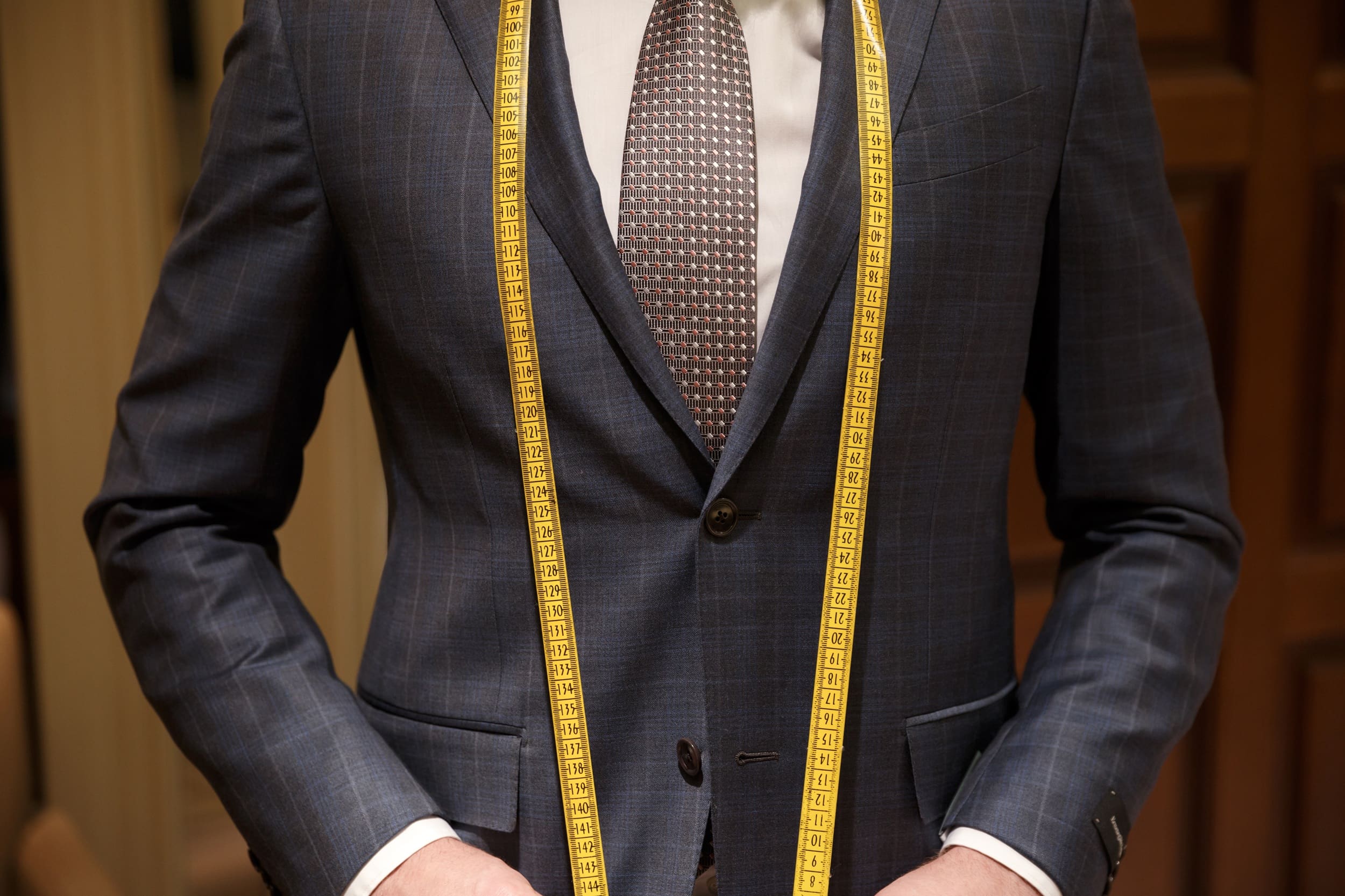 A person wearing a blue checkered suit and tie stands with a yellow measuring tape draped around their neck.
