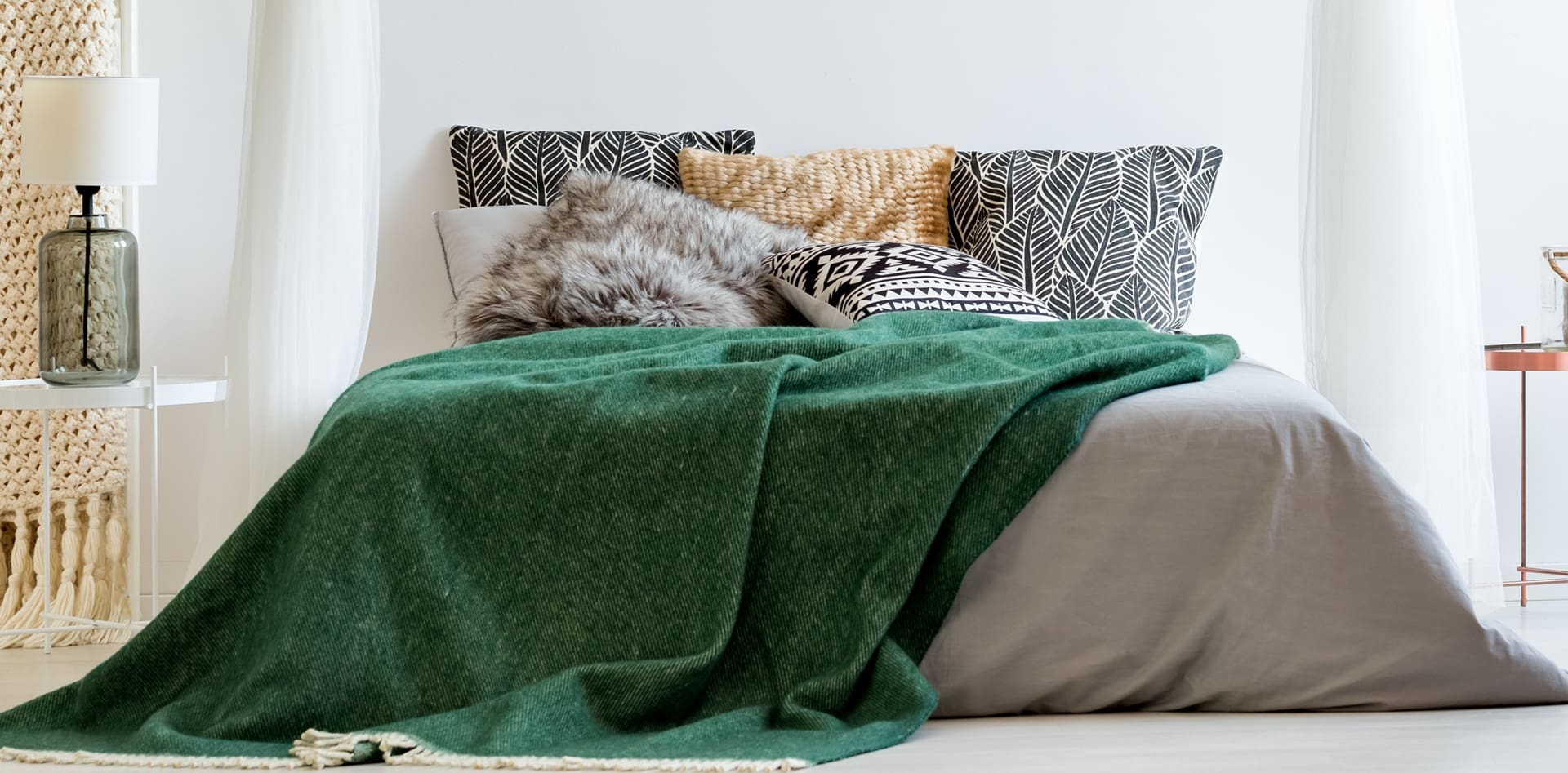 A neatly made bed with grey sheets, assorted patterned pillows, and a green blanket in a modern, well-lit bedroom.