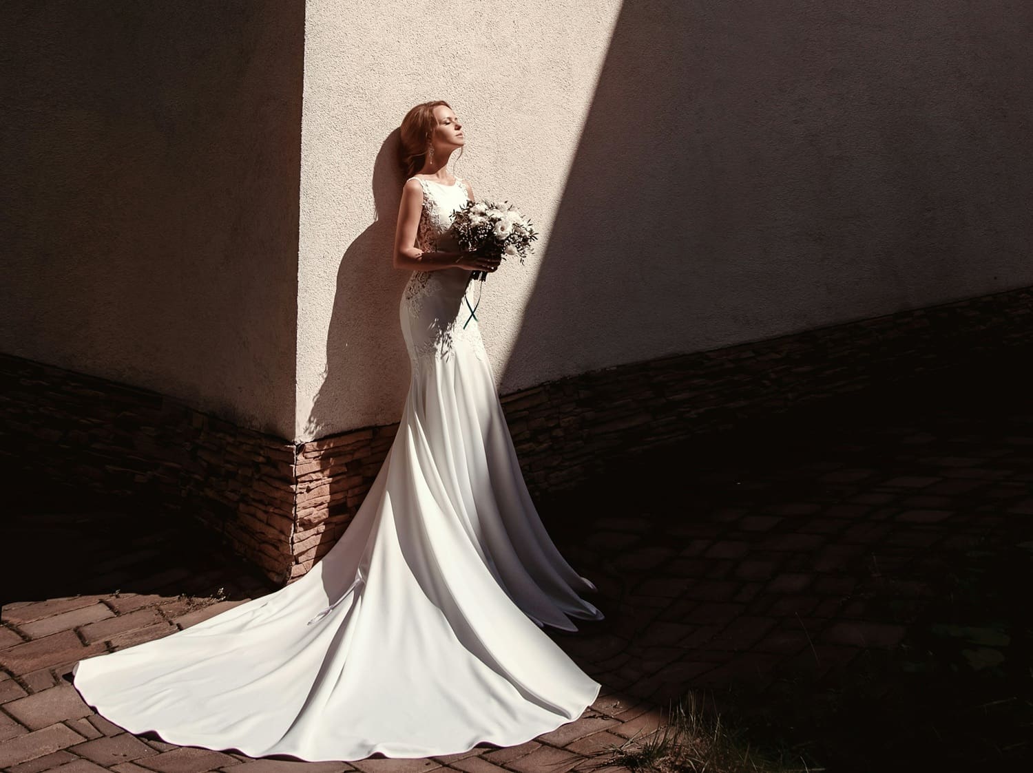 Bride in elegant wedding dress with train, holding bouquet, standing against a textured wall, emphasizing wedding dress preservation and care.