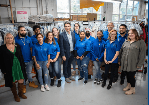 Group photo of Best Cleaners team members in blue uniforms, standing with a smiling man in a blazer, surrounded by garment bags, celebrating the transition from Feigenbaum Cleaners to Best Cleaners NY.