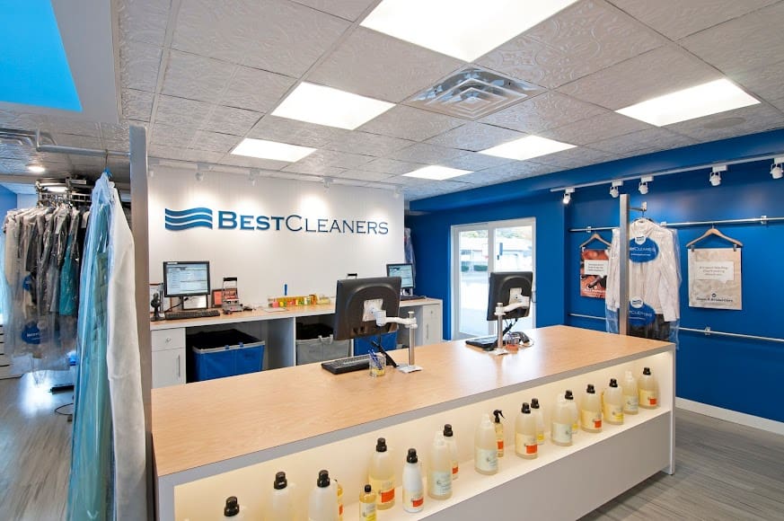 Modern dry cleaning shop interior with a wooden counter, computer stations, cleaning product bottles, and garments hanging behind the counter.