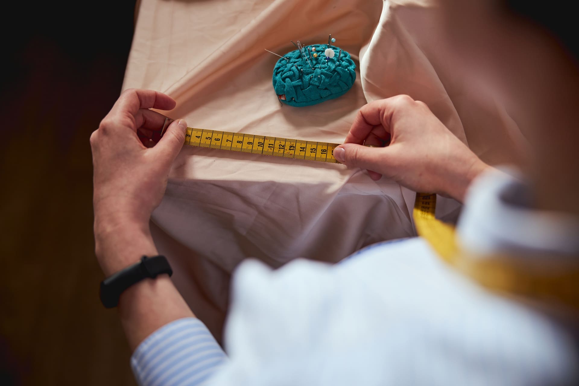 Person measuring fabric with a yellow measuring tape; a blue pincushion with pins is visible on the fabric.
