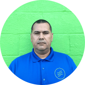 A man in a blue collared shirt stands against a bright green brick wall, facing the camera with a neutral expression.