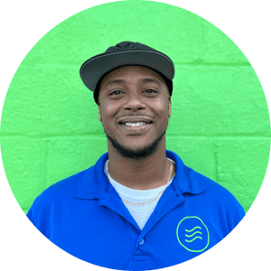 A man wearing a black cap and blue polo shirt stands smiling in front of a bright green painted brick wall.