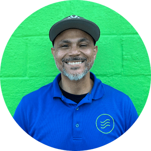 A man wearing a blue collared shirt and a gray cap stands in front of a bright green brick wall, smiling at the camera.