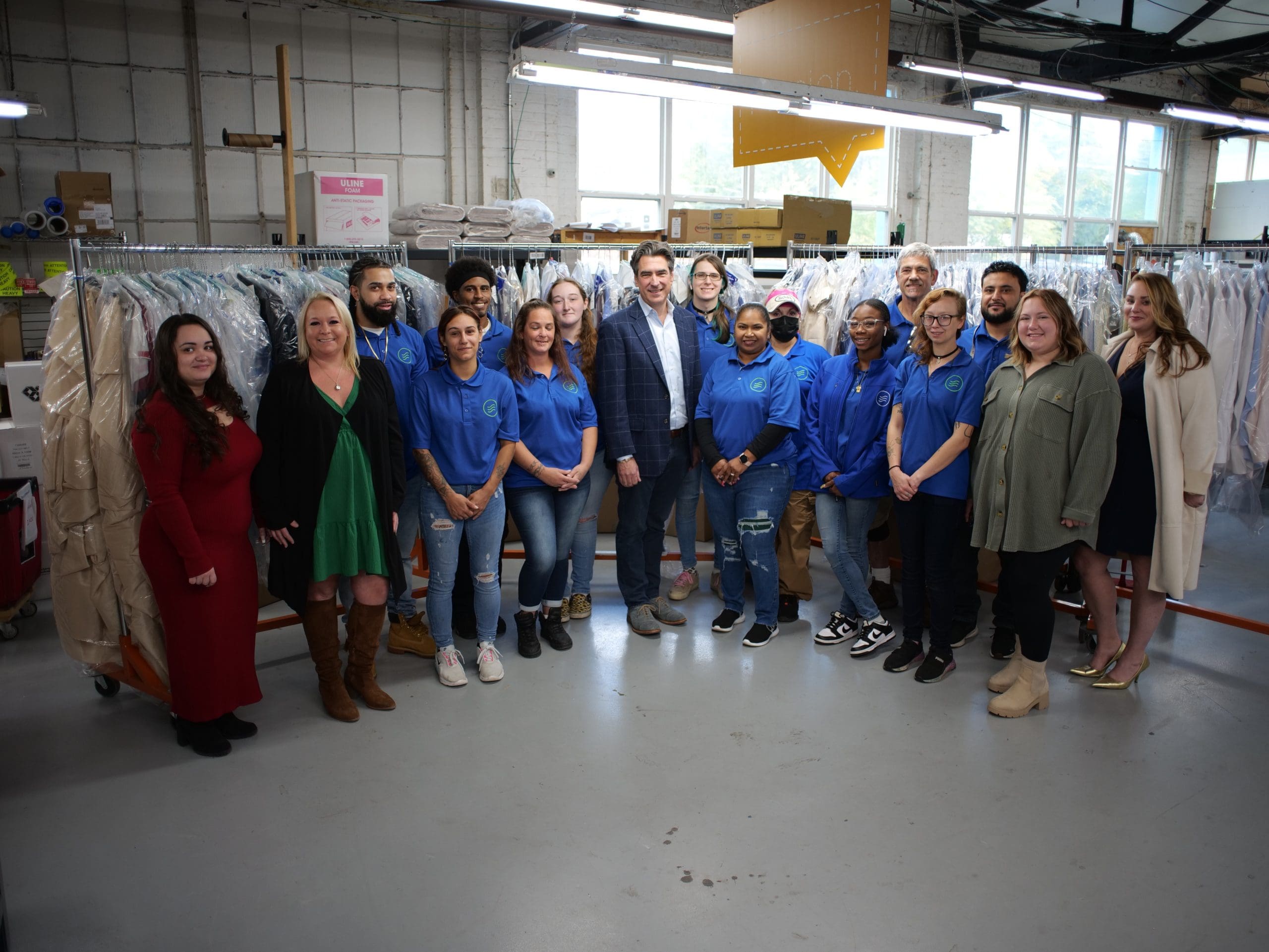 A group of people stand together in a dry cleaning facility, with clothing racks and packaged garments visible in the background.
