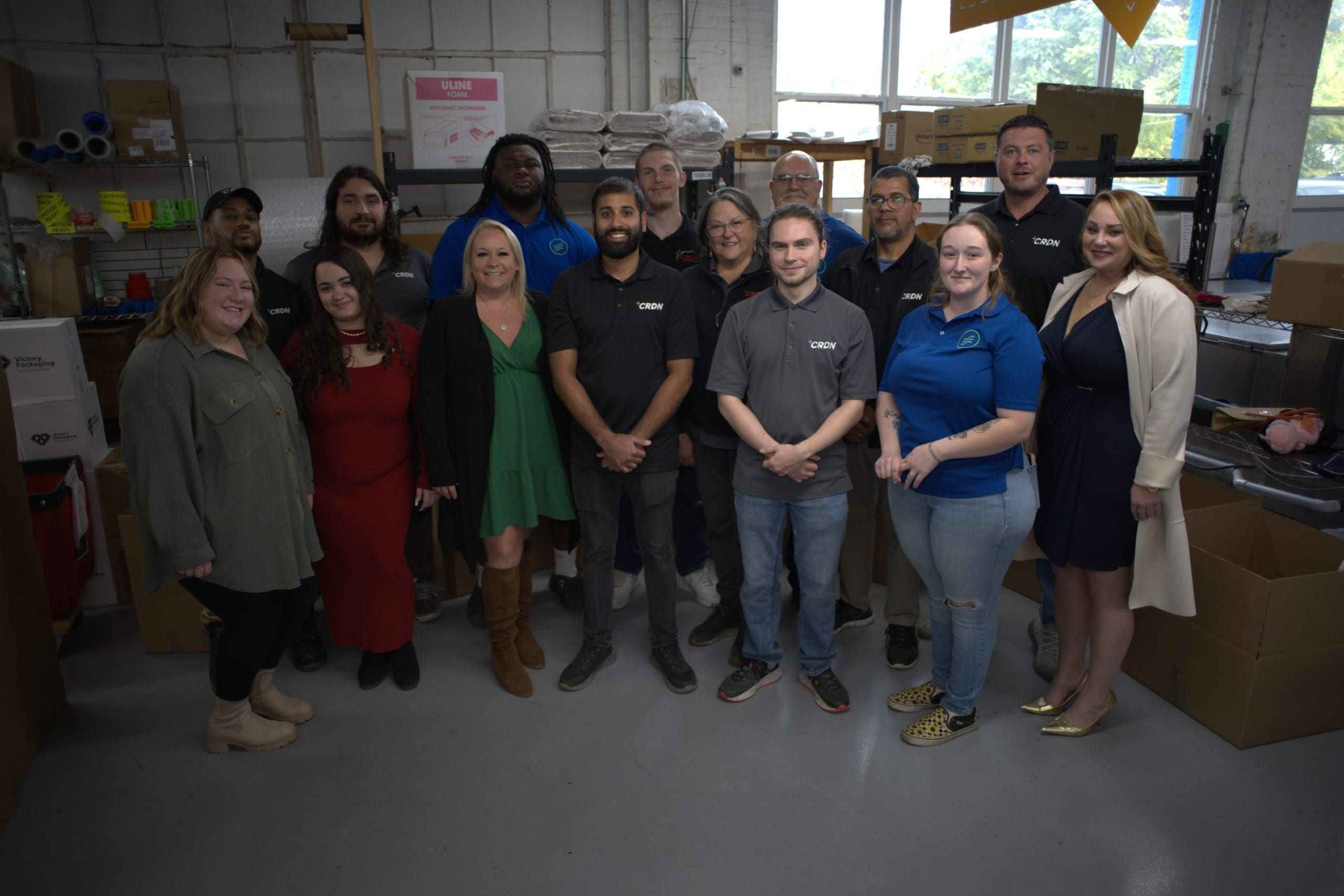 A group of fifteen people stand together in a warehouse-like workspace, posing for a group photo. Boxes and industrial supplies are visible in the background.