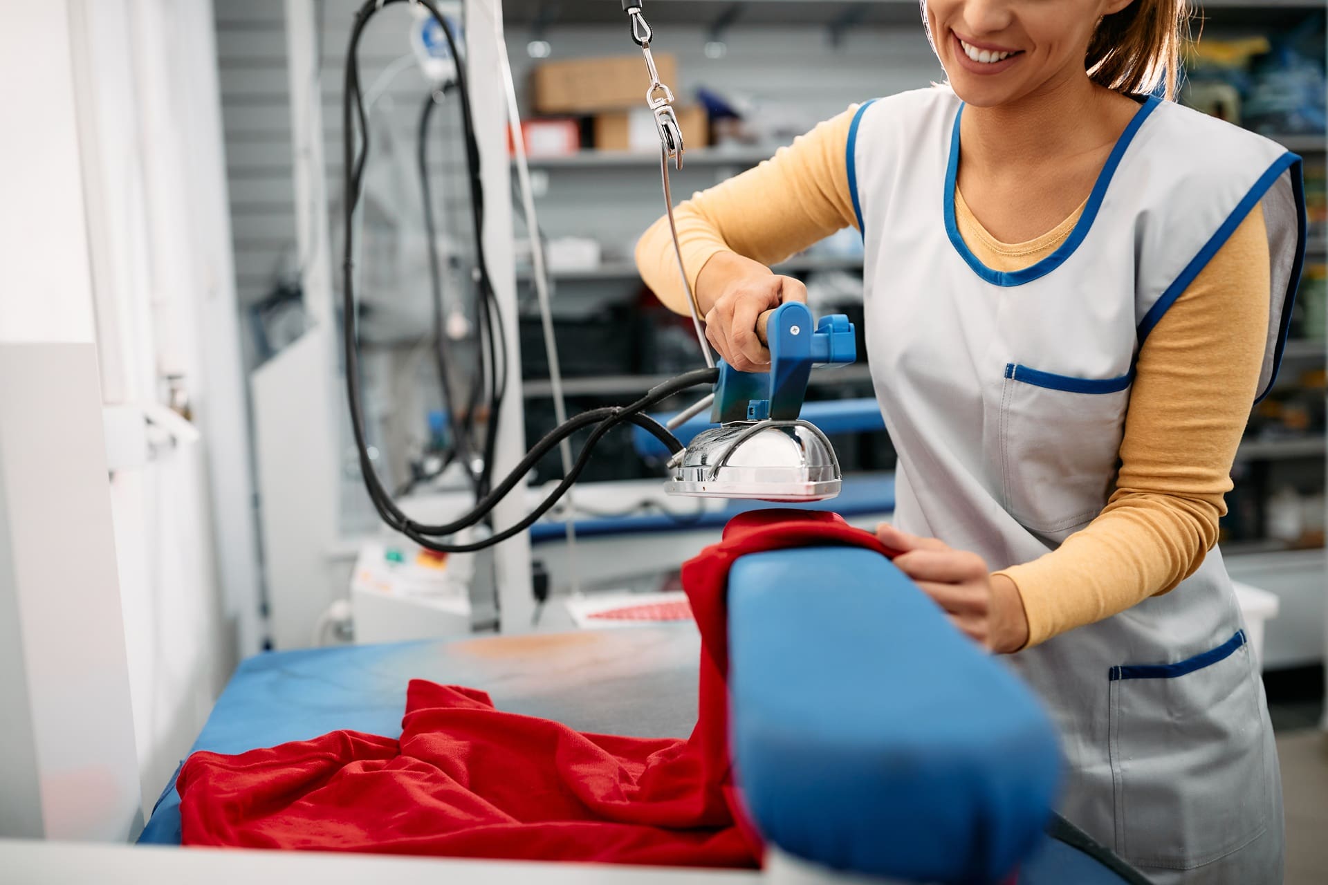 A person in a uniform ironing a red garment on a blue ironing board in a laundry or dry cleaning facility.