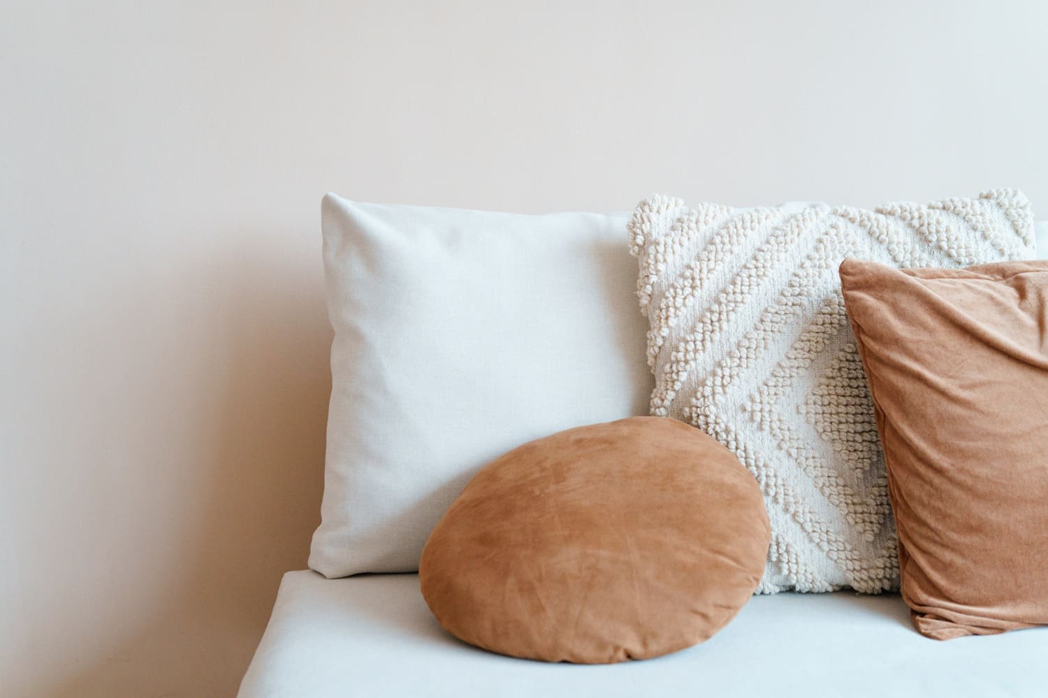 A light-colored sofa with three decorative pillows: one square beige, one textured cream, and one round tan pillow.