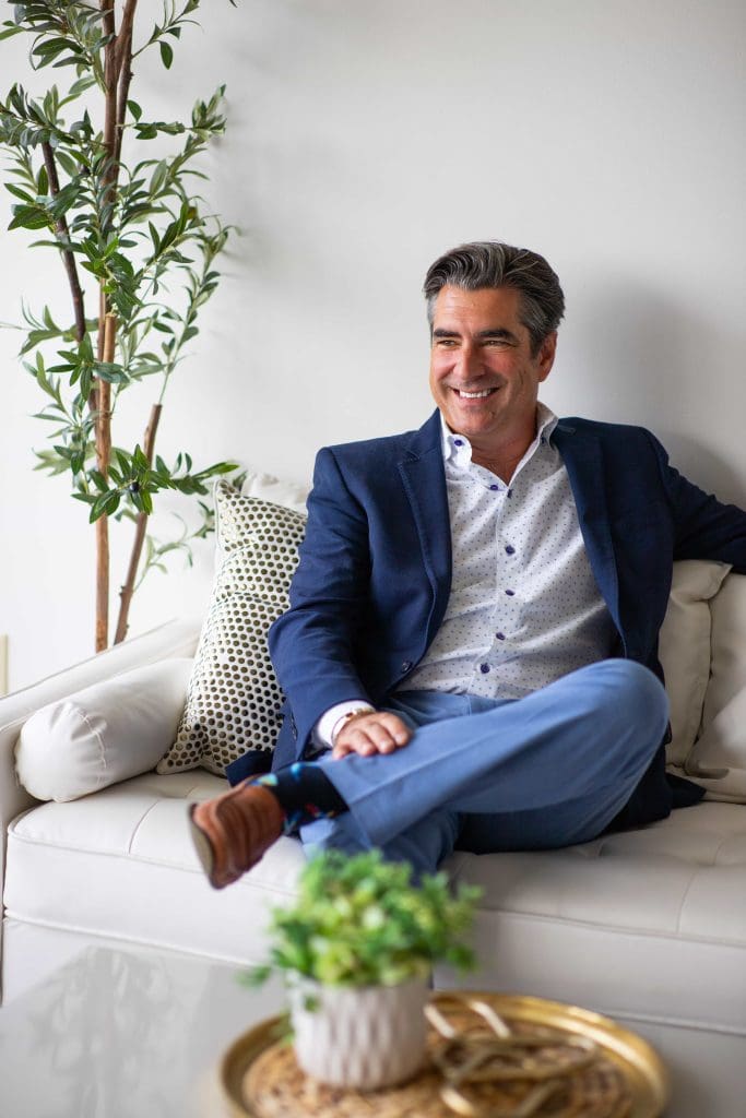 A man in a blue suit jacket and light blue pants sits on a white sofa, smiling, with a green plant and gold tray visible in the foreground.