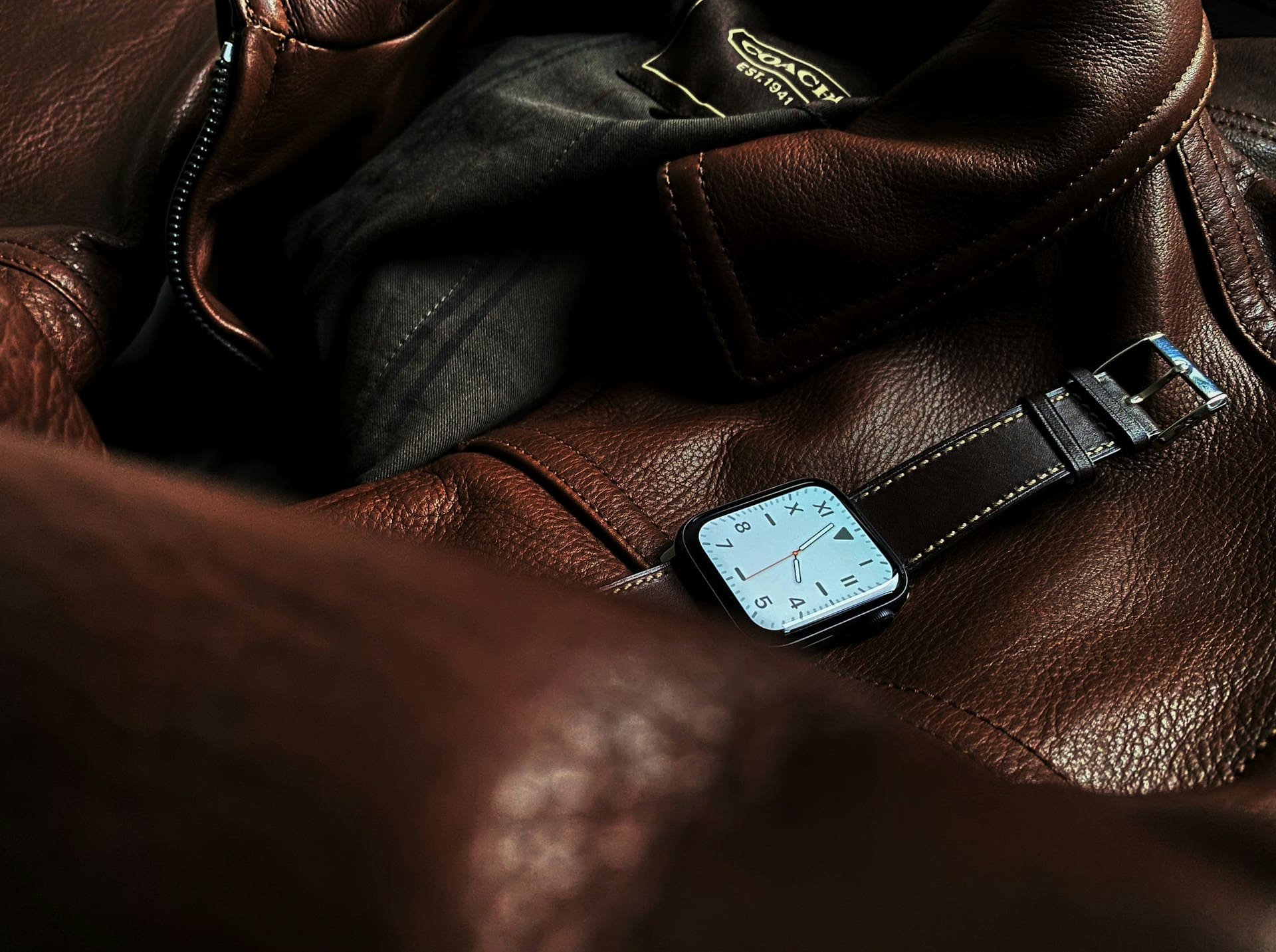A brown leather strap smartwatch rests on a dark brown leather jacket with a visible Coach label in the background.