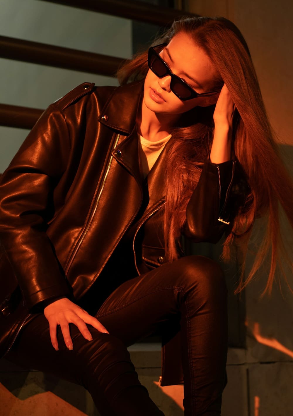 A person wearing a black leather jacket, black sunglasses, and black pants sits on a step, touching their hair with one hand.