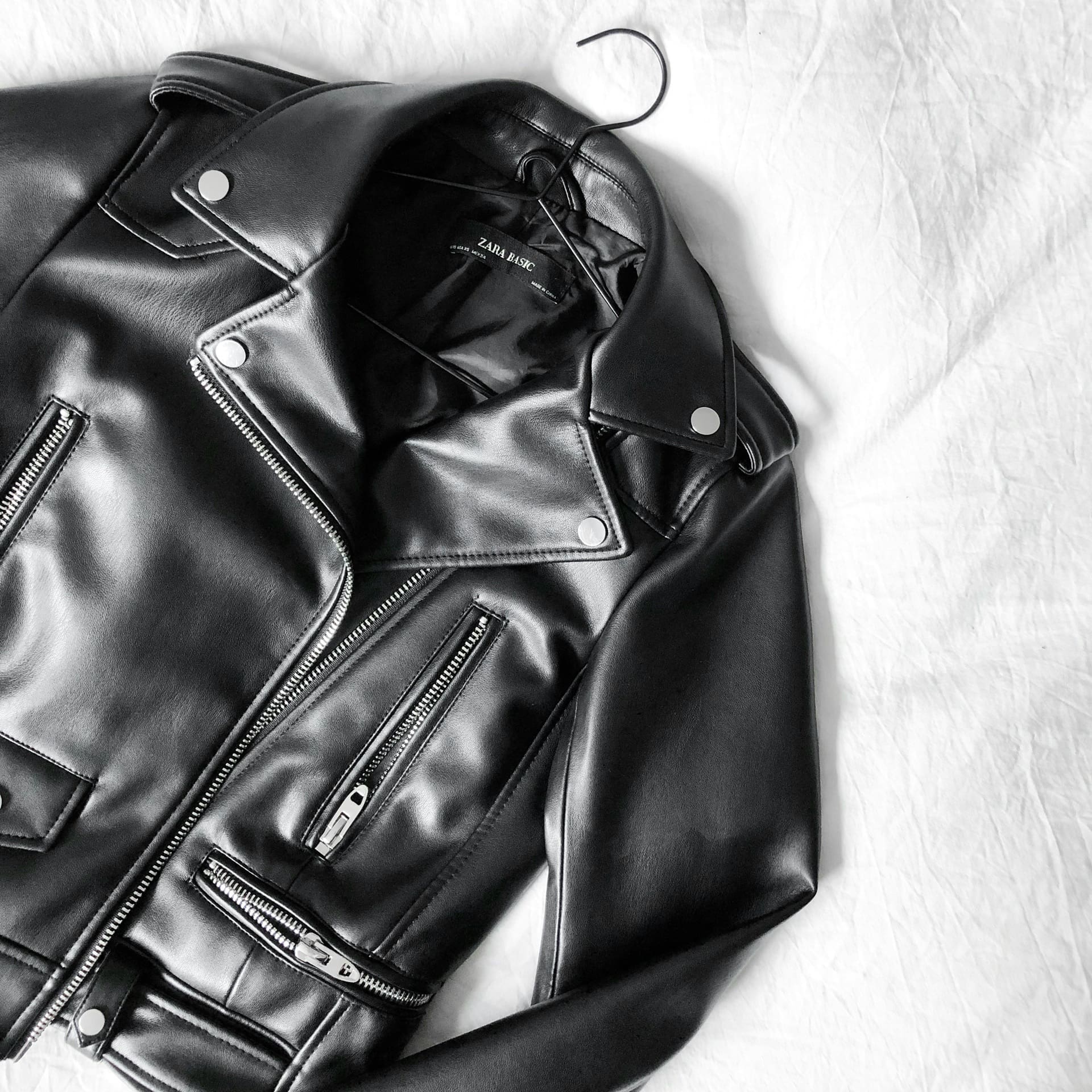 A black faux leather biker jacket with silver zippers and buttons is laid flat on a white textured surface.