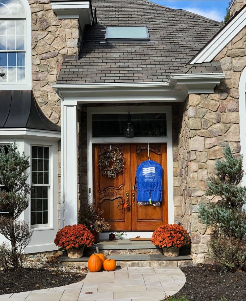 Stone house with double wooden front doors, autumn wreaths, potted mums, and two pumpkins on the steps; a blue jacket hangs on one door.