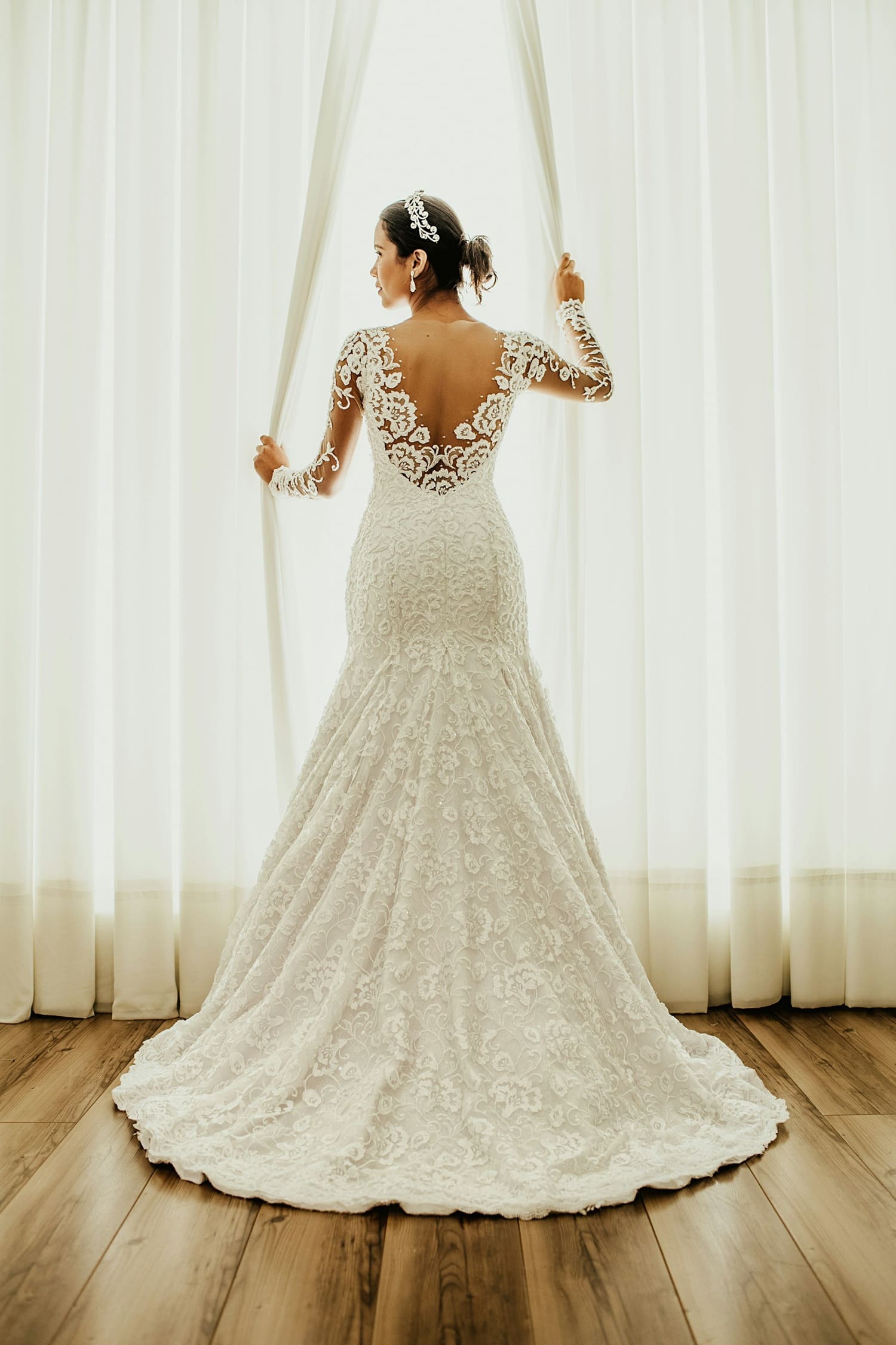 Bride in intricate lace wedding dress with detailed embellishments, standing by sheer curtains, highlighting expert craftsmanship and preservation importance for wedding gowns.