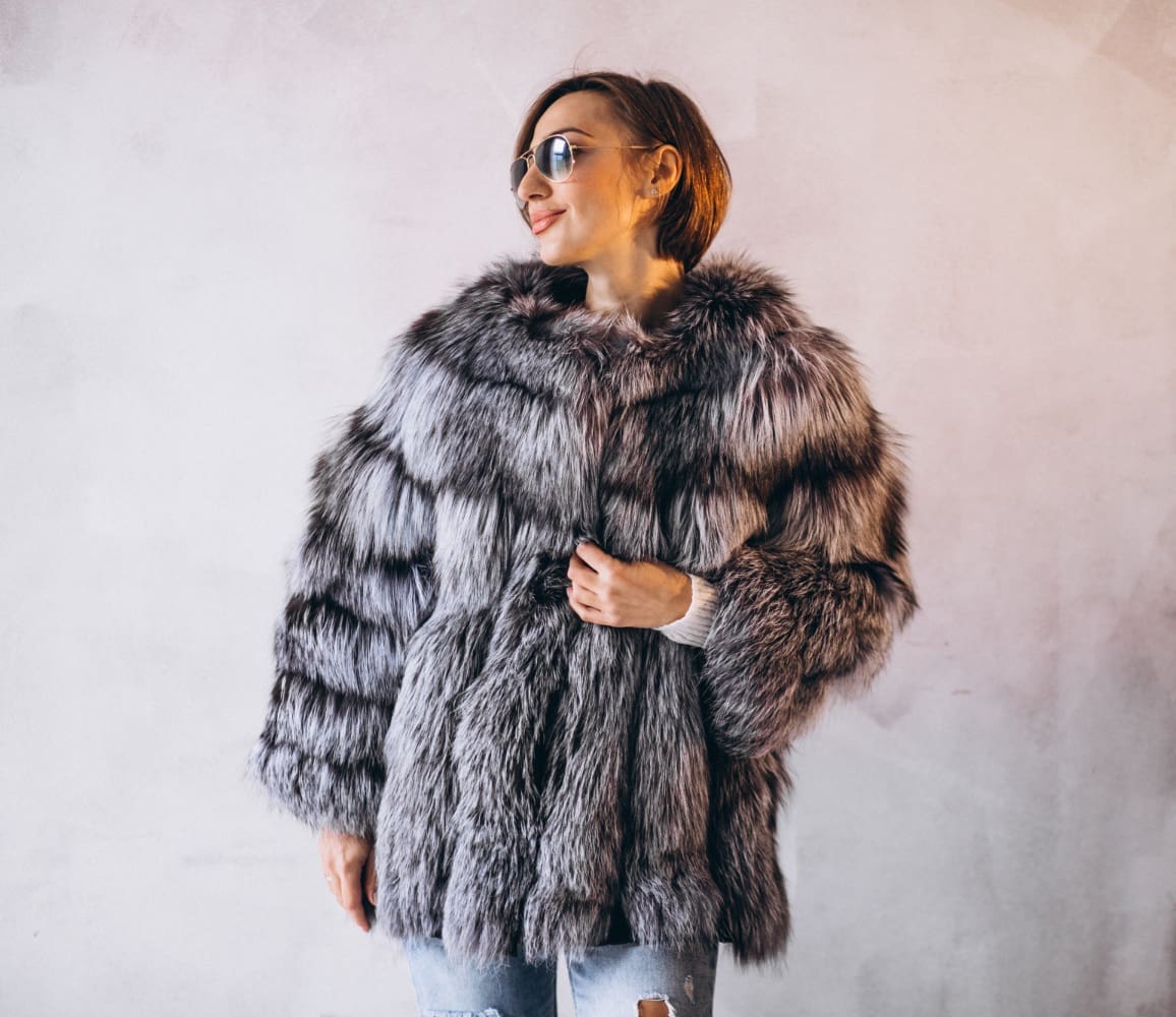 Person wearing sunglasses and a large, fluffy gray fur coat stands against a plain light-colored wall, with one hand holding the coat closed.