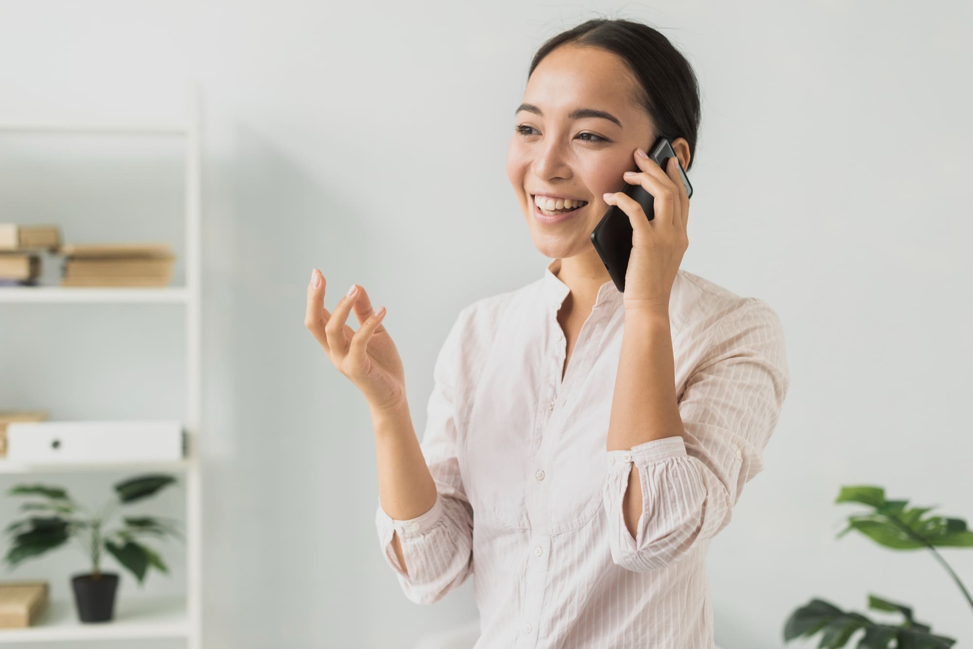 A woman in a white shirt smiles and gestures with her hand while talking on a mobile phone in a bright office setting.