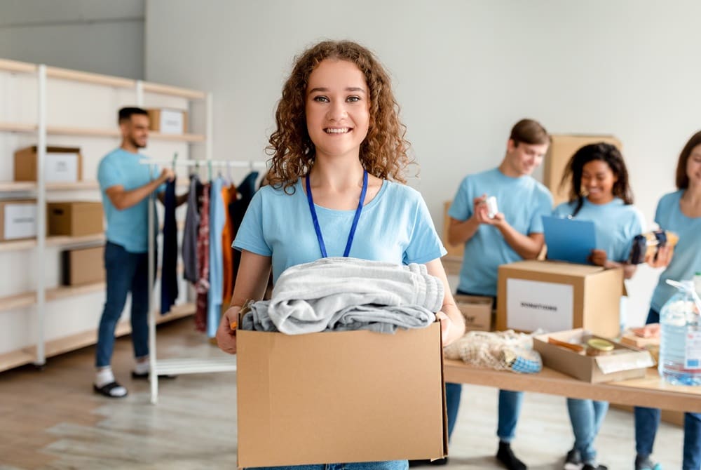 Young woman holding a donation box with folded clothes, standing in front of a group of people sorting items in a donation center.