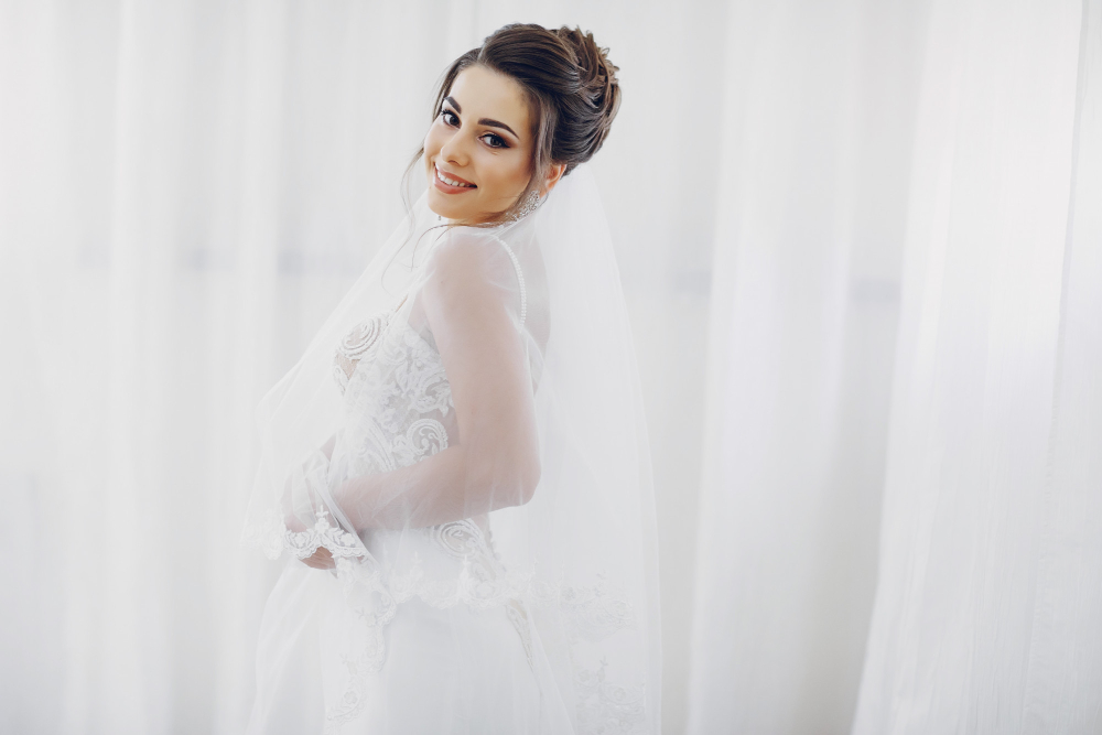 Bride in elegant wedding dress with veil, smiling in front of sheer white curtains, representing wedding dress care and preservation.