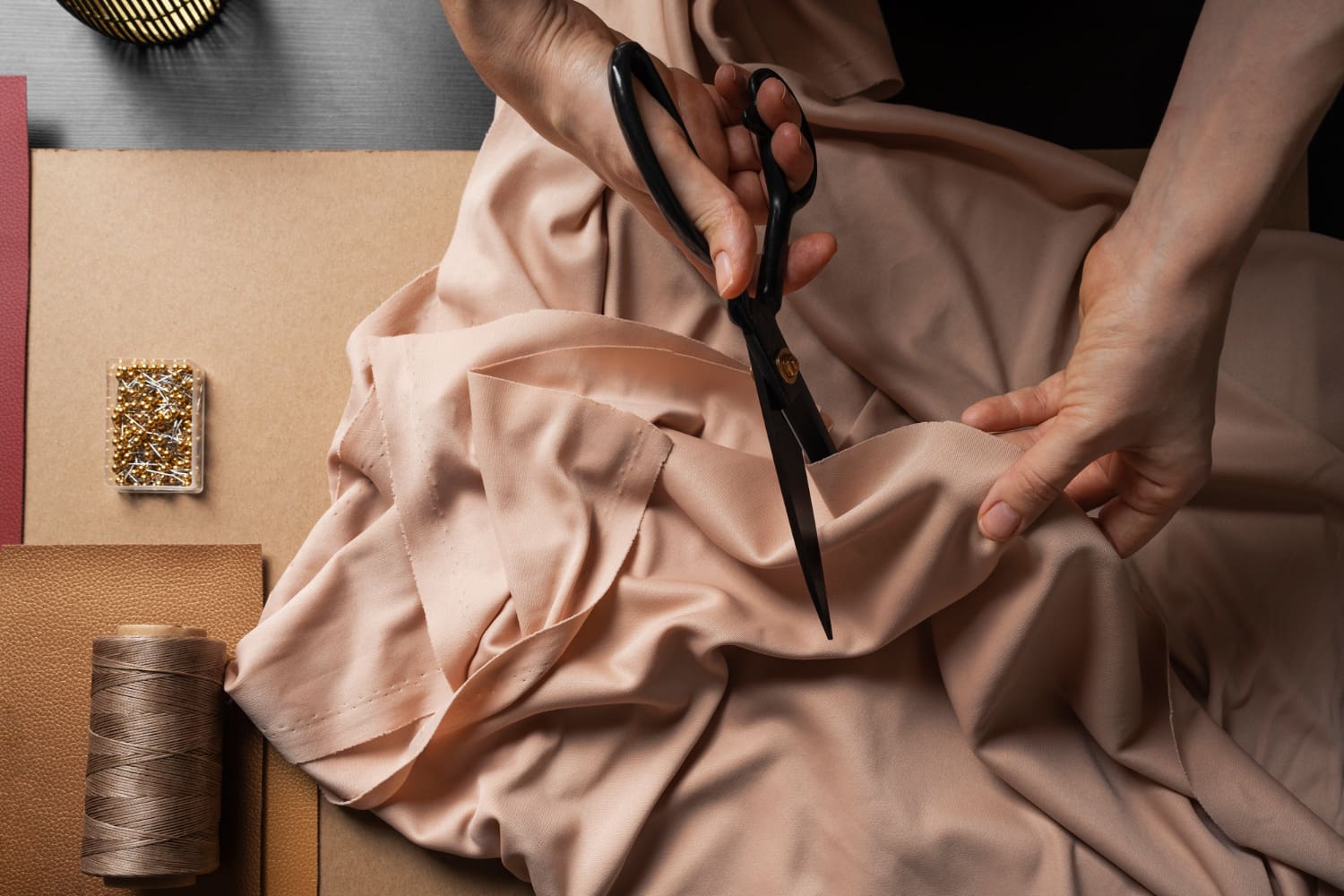 A person cuts light pink fabric with black scissors on a table with thread, pins, and other sewing materials.