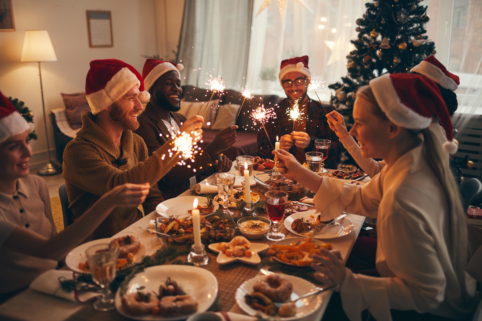 A group of people wearing Santa hats sit around a festive dinner table, holding sparklers and celebrating together in a decorated room with a Christmas tree.