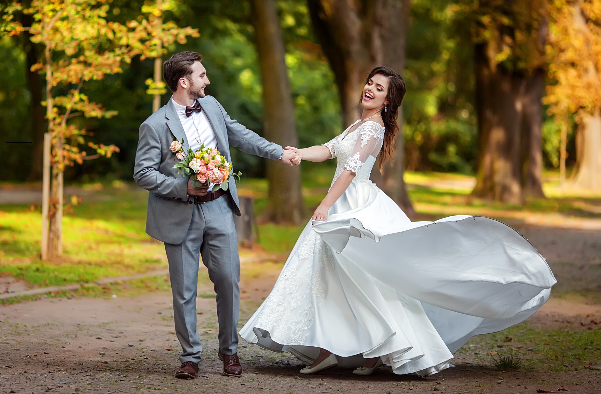 A bride and groom hold hands and smile as the bride twirls in her white dress outdoors on a tree-lined path.