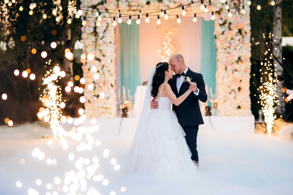 A bride and groom share their first dance outdoors, surrounded by flowers, string lights, and pyrotechnic effects.