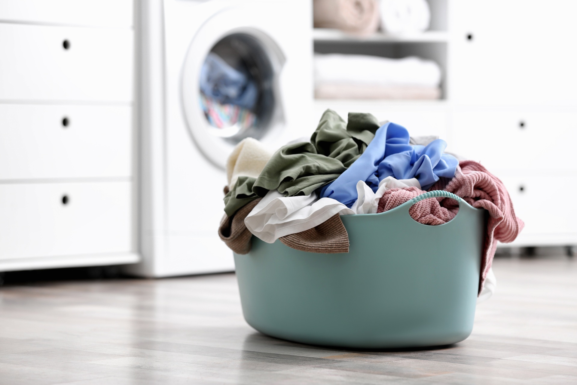 A full laundry basket filled with assorted clothes sits on the floor in front of a washing machine in a laundry room.