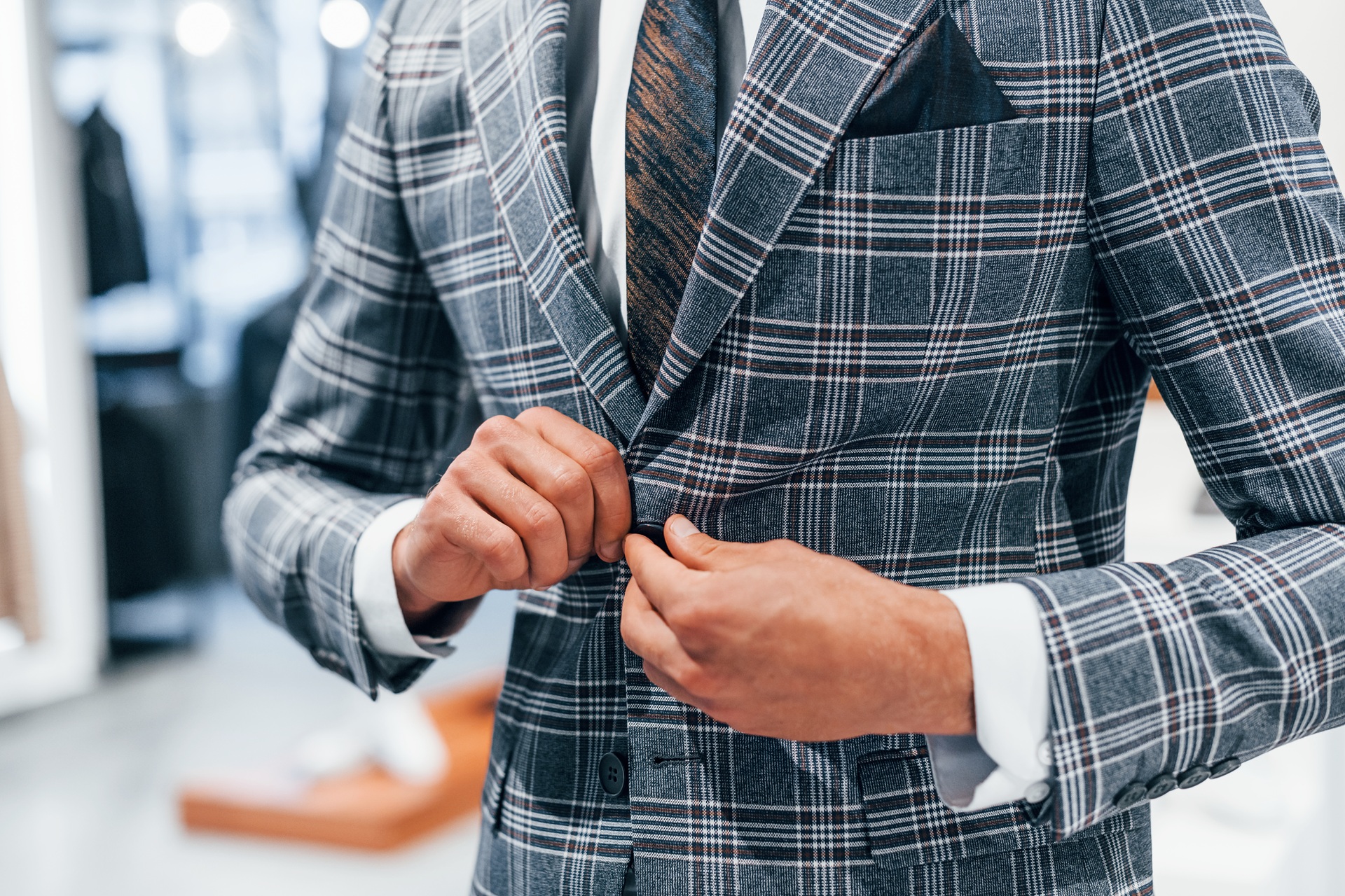 Man in a plaid suit jacket buttoning the front, with a white dress shirt, patterned tie, and pocket square visible.