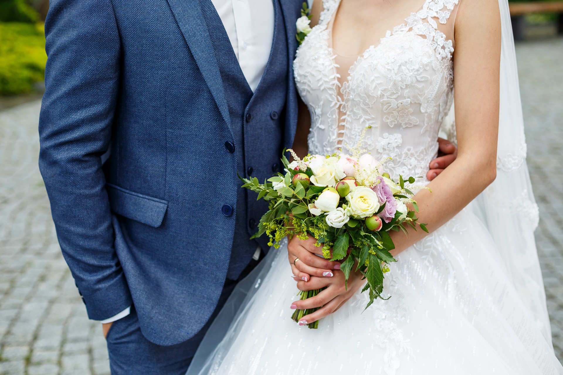 Bride in a white lace wedding dress holds a bouquet of flowers while standing next to a groom in a blue suit on a cobblestone path. Their faces are not visible.