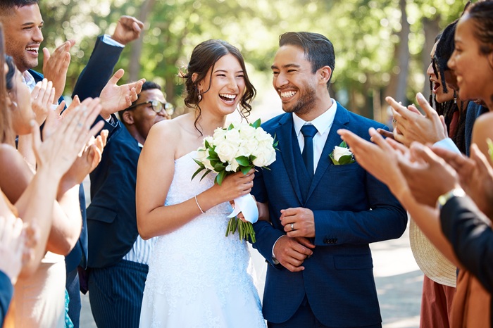 Bride and groom smiling and holding flowers, surrounded by friends clapping and celebrating outdoors on a sunny day.
