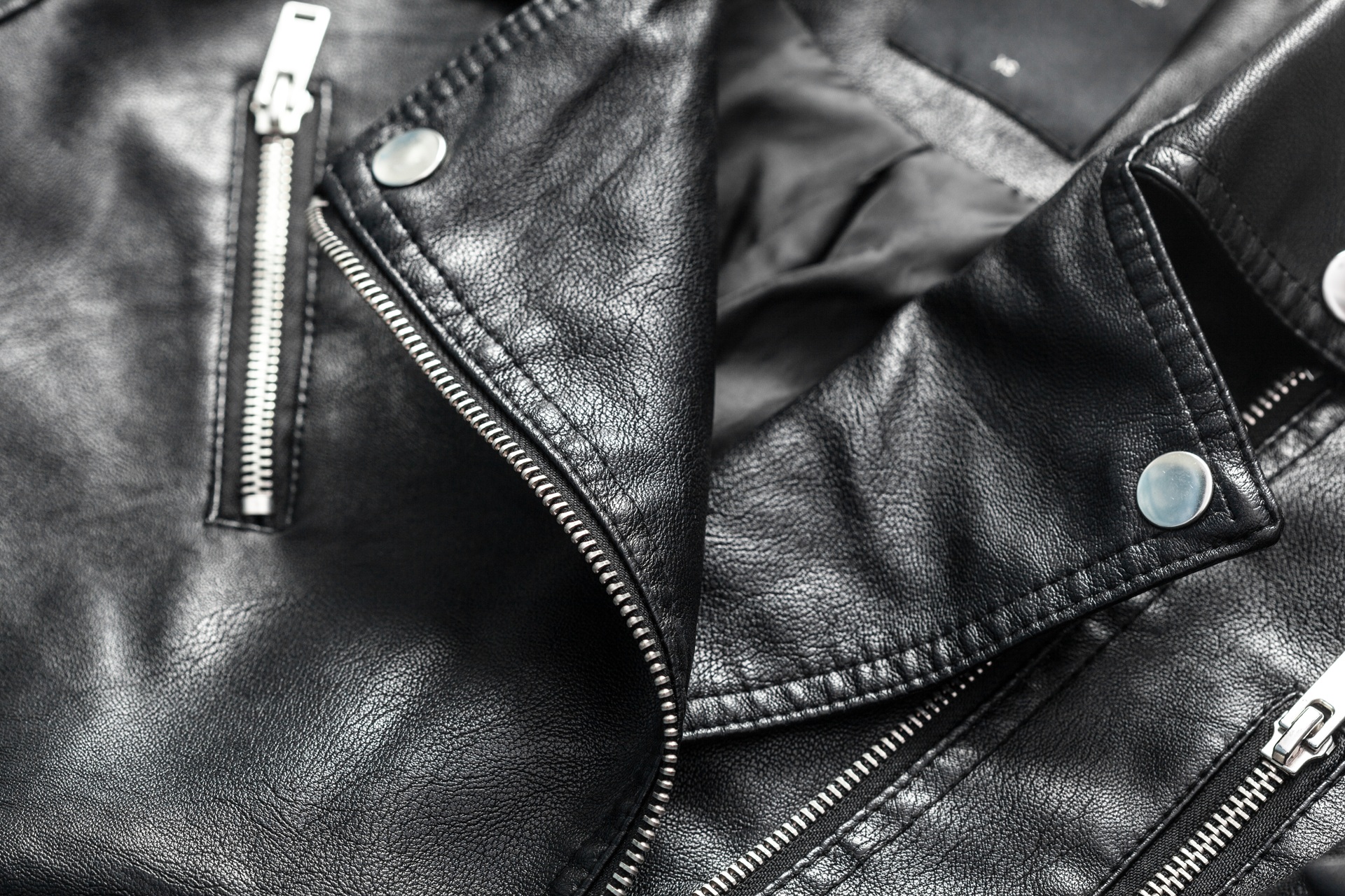 Close-up of a black leather jacket showing silver zippers, snap buttons, and textured material with visible stitching.