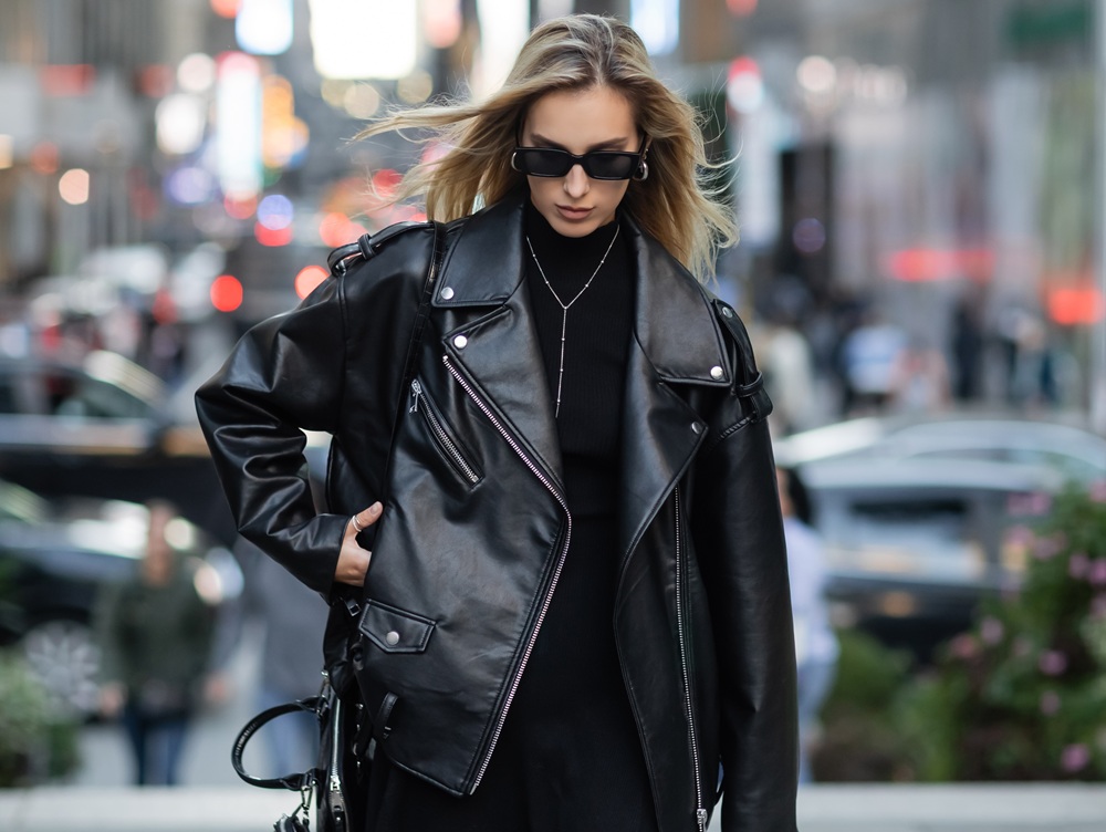 A woman wearing a black leather jacket, black sunglasses, and a black outfit walks on a city street with blurred cars and lights in the background.