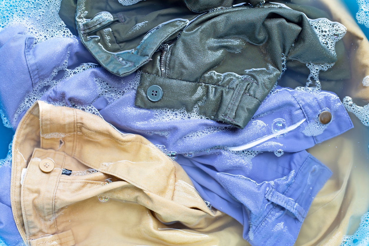 Four pairs of colored pants are soaking in soapy water, partially submerged, with visible bubbles and buttons.